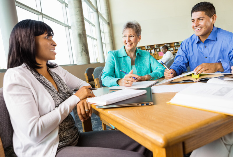 Three coworkers sitting around table in library working together communicating writing notes. 