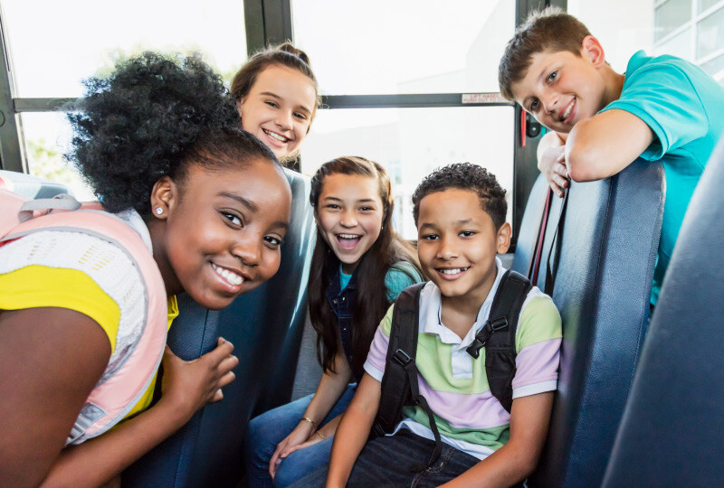 Five students sitting on school bus carrying backpacks smiling. 