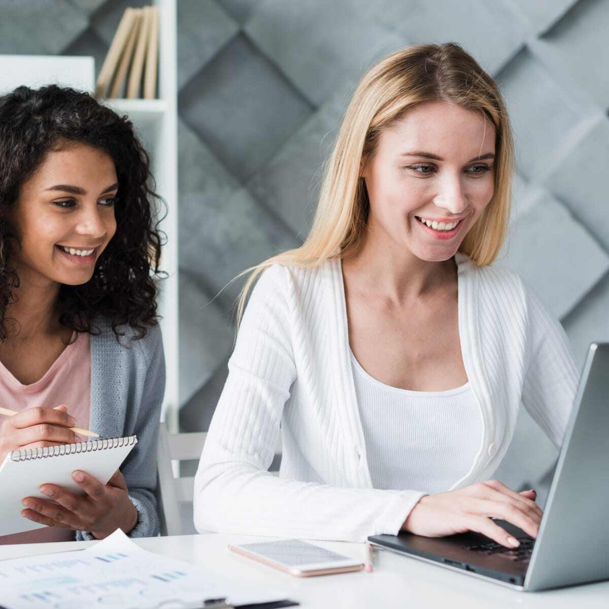 Two women smiling working together at desk laptop pen and paper. 