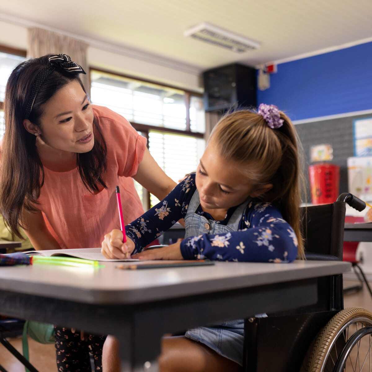 Teacher helping student girl in wheelchair at desk in classroom. 