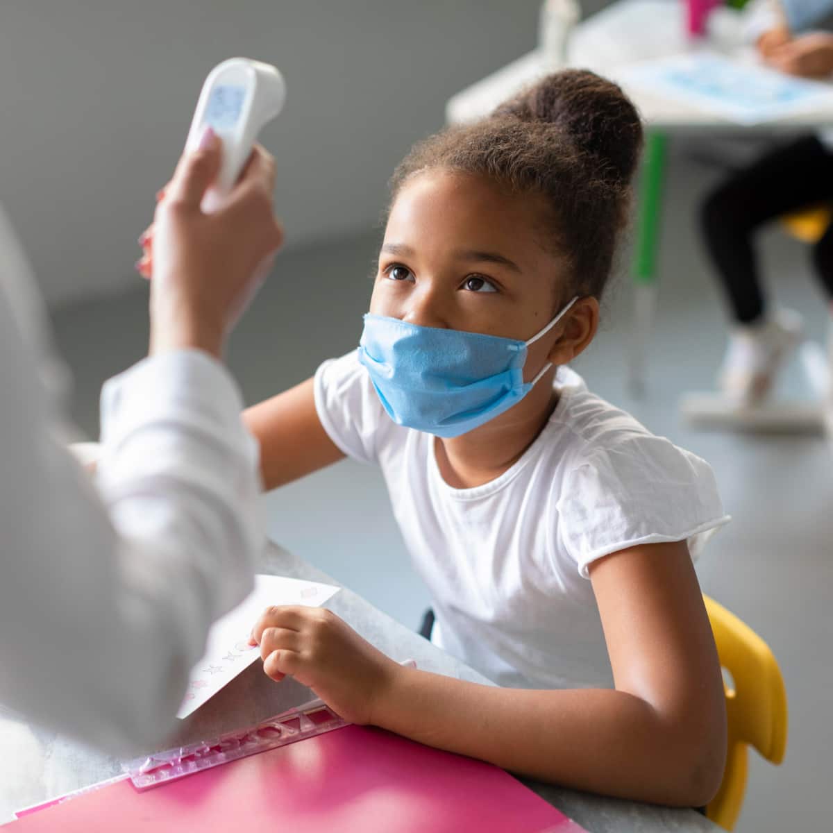 School nurse taking temperature of student sitting at desk in classroom. 