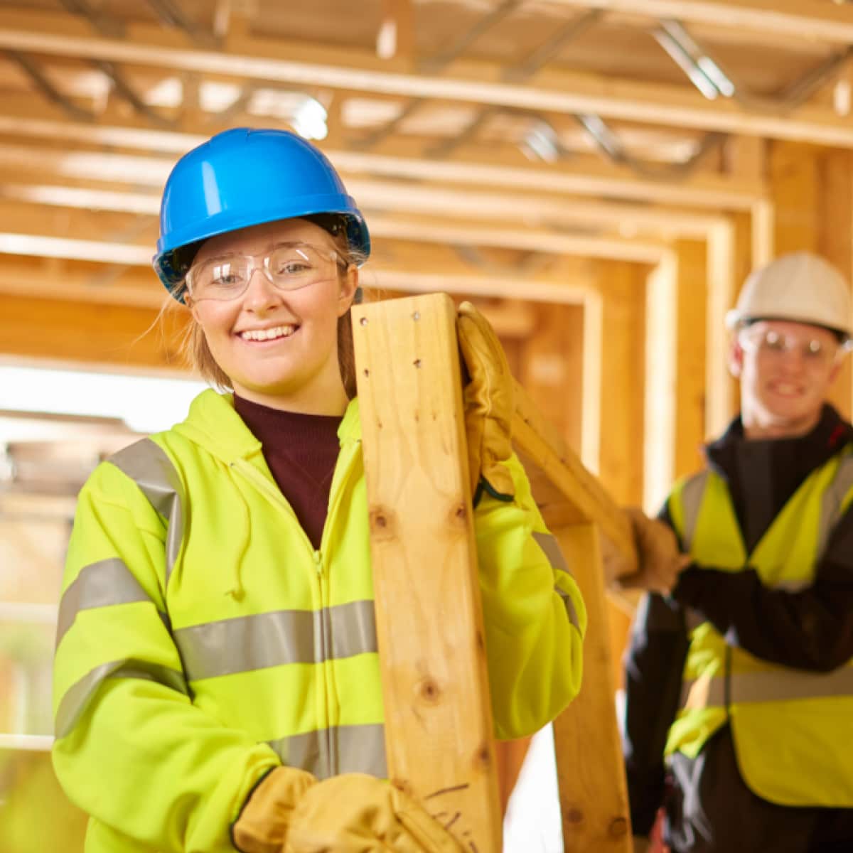 Woman and man smiling facing forward wearing safety goggles and vests working in construction area. 