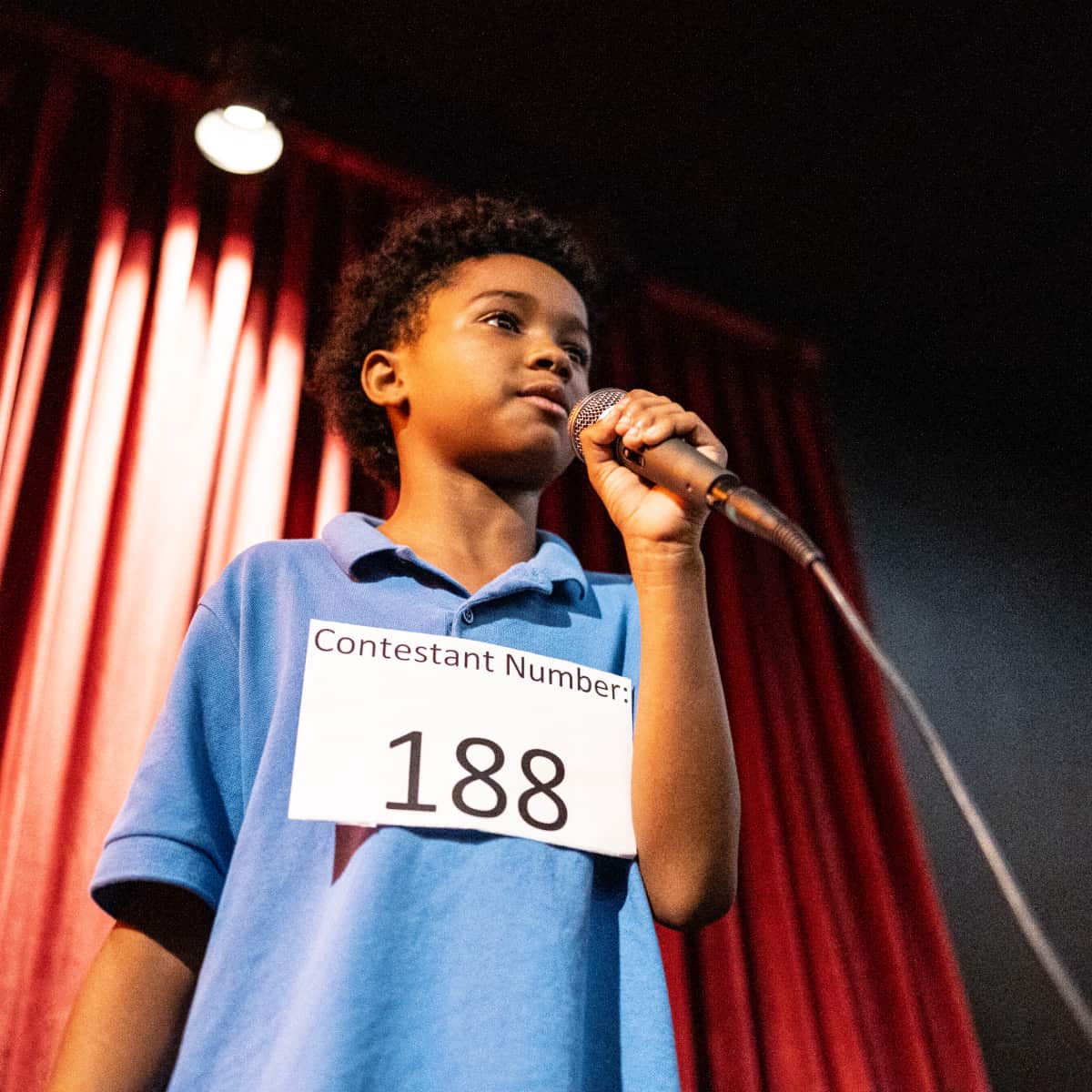 Student speaking intro microphone on stage in front of red curtain for spelling be competition. 