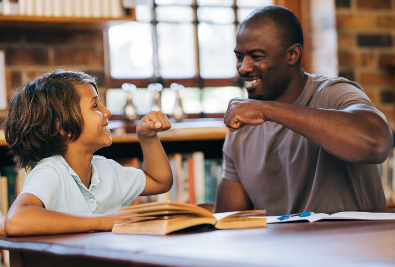 Man and young boy sitting at table facing each other smiling and fist bumping. 