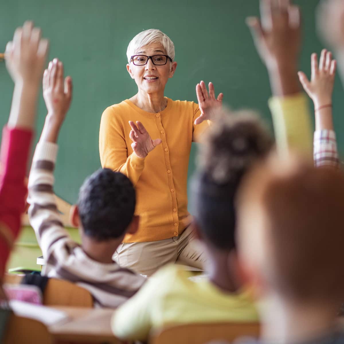 Teacher sitting in front of classroom students at desks listening and raising hands. 