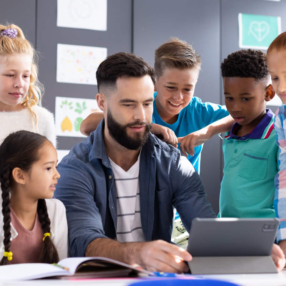 Six students standing around teacher at table looking at tablet learning. 