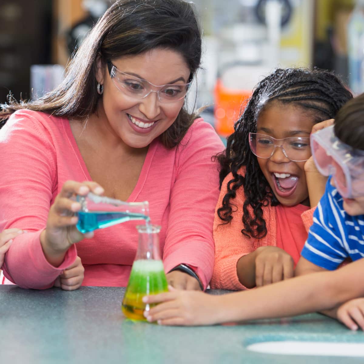 Four students and teacher sitting around desk during science wearing goggles doing experiment. 