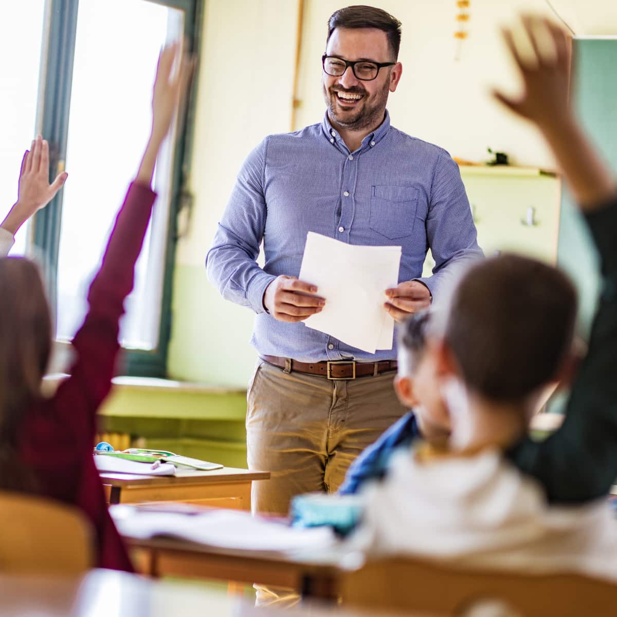 Students in classroom raising hands male teacher holding papers smiling and teaching. 