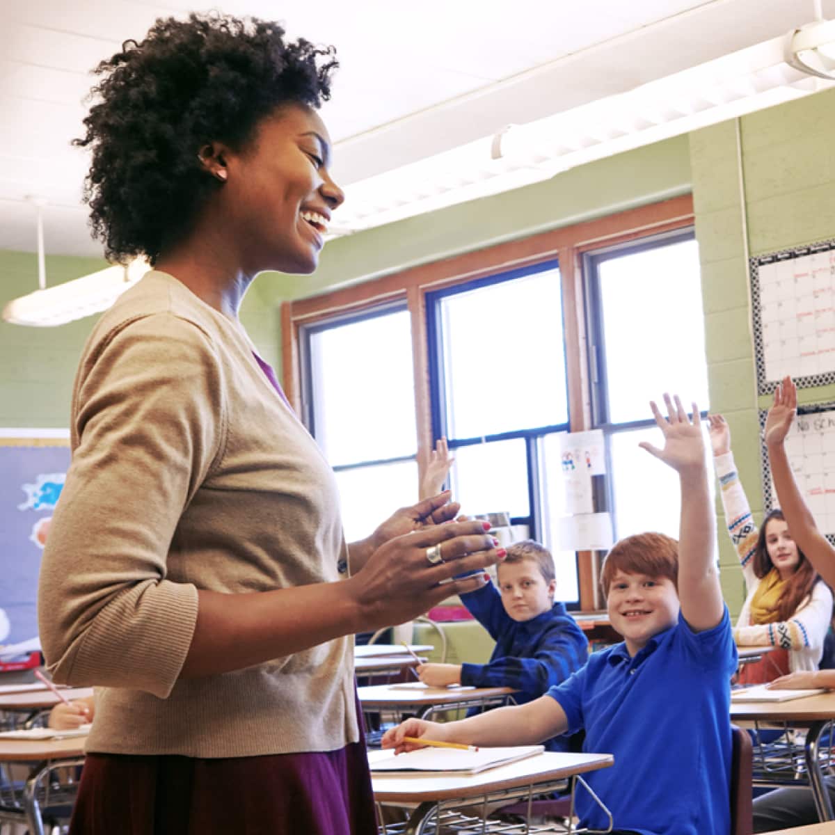 Teacher smiling standing front of classroom facing students sitting at desks raising their hands.