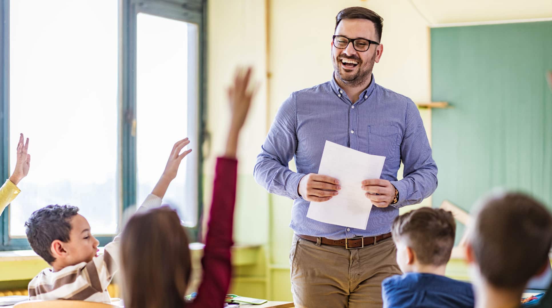 Teacher standing in front of classroom smiling facing students who are sitting in desks raising their hands. 