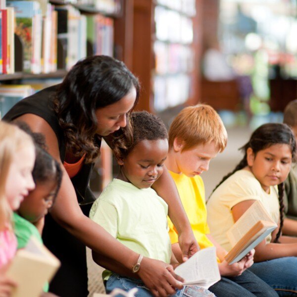 seven students sitting in library reading books teacher helping one student