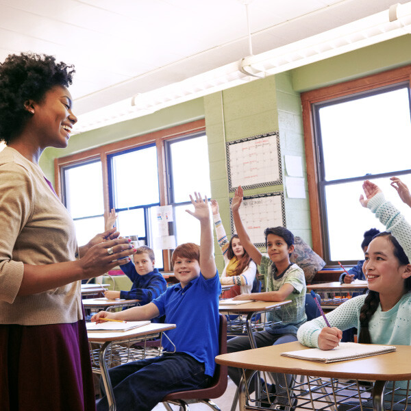 teacher standing front of classroom looking at students sitting in desks raising hands