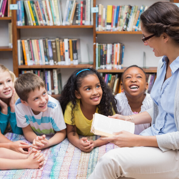 six students sitting on floor in library teacher sitting next to them reading them a book