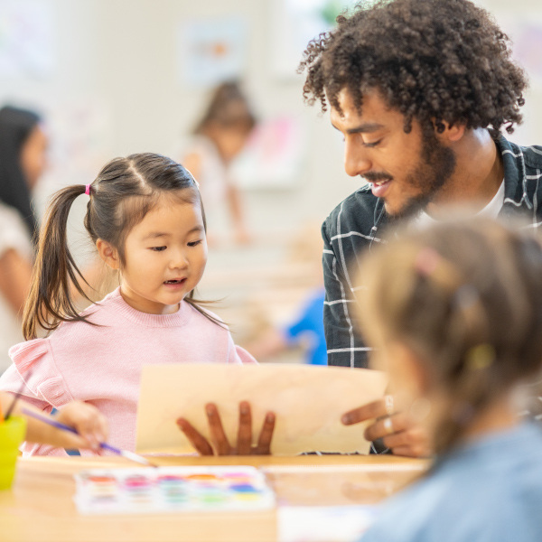 students sitting at table in classroom painting teacher sitting next to them helping