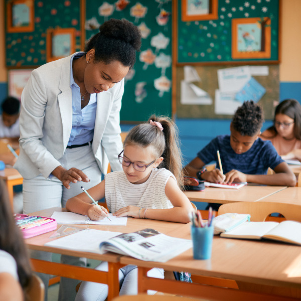 students sitting at desks in classroom writing in notebooks teacher standing helping