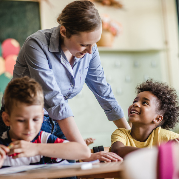 young students sitting at desk in classroom teacher standing behind helping with assignment