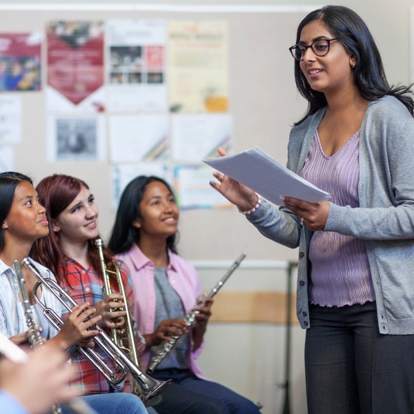 students sitting next to each other holding musical instruments teacher standing facing them talking 