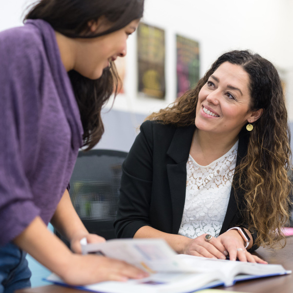 teacher sitting at desk student standing asking question from workbook smiling