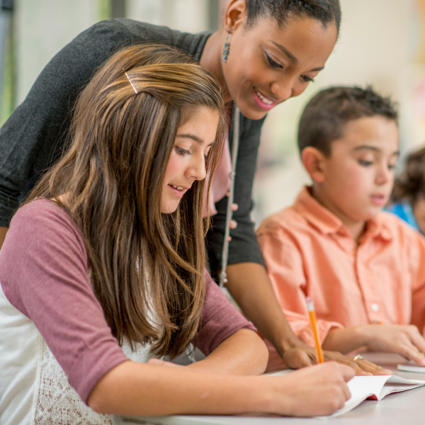 students sitting at desk in classroom writing in notebooks teacher standing helping 