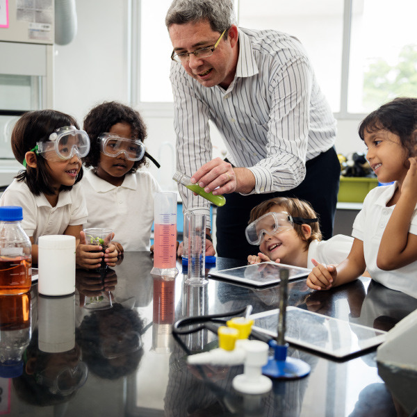 students and teacher wearing safety goggles sitting around table doing a science experiment