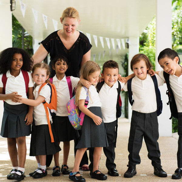 young students and teacher standing in line next to each other facing forward smiling holding backpacks