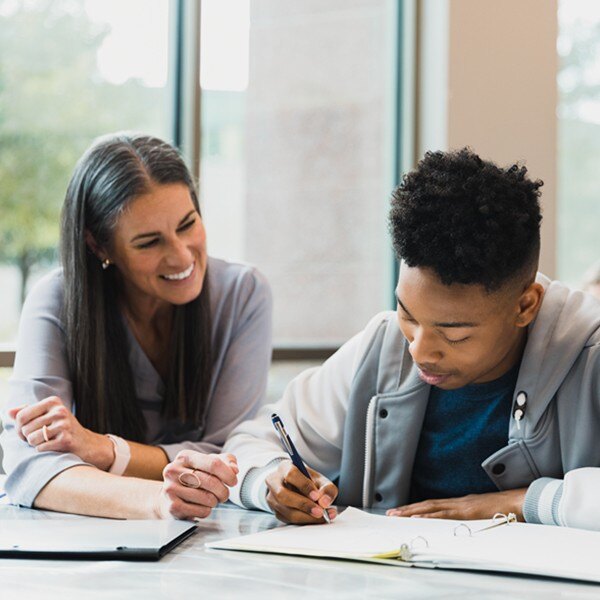 teacher helping student with assignment sitting at desk with pens and binder