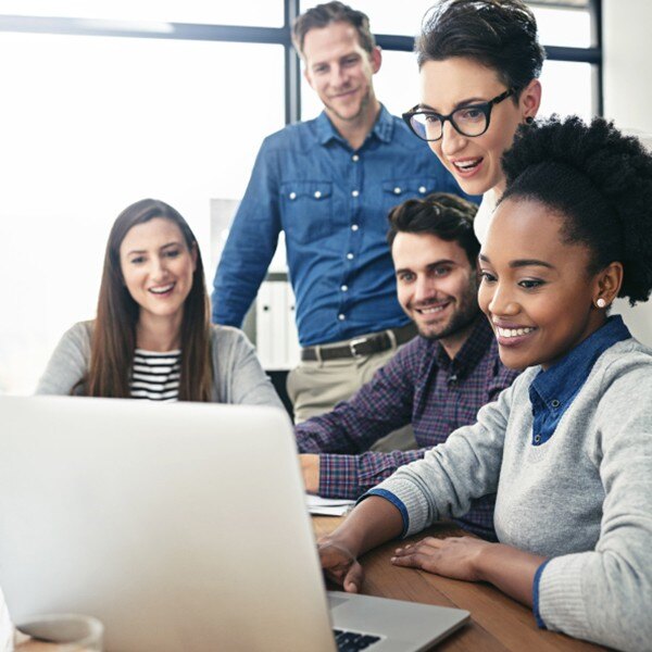 five coworkers sitting around desk working together smiling looking at laptop