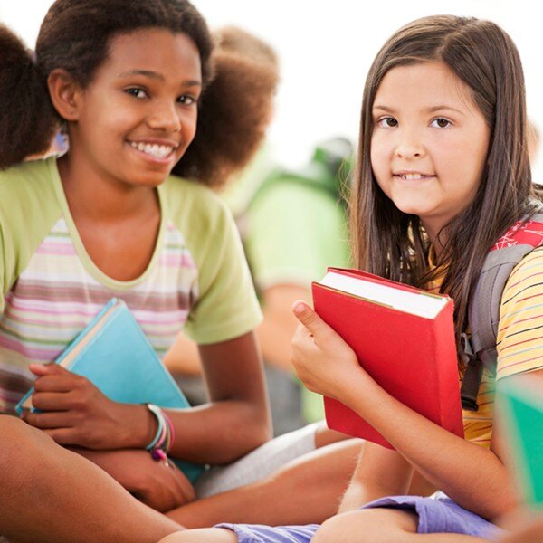 children sitting on the floor facing forward smiling carrying a backpack and holding books