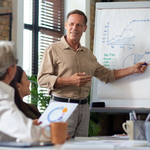 two coworkers sitting at table in conference room one coworker standing pointing at data chart