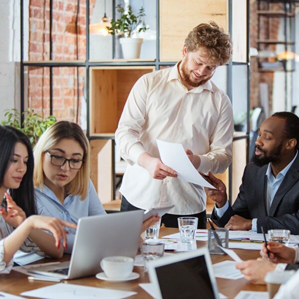 five coworkers sitting and standing around desk in conference room talking working together looking at documents and laptop