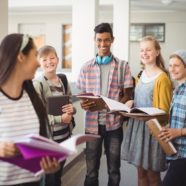 five students standing in school hallway carrying backpacks and books smiling