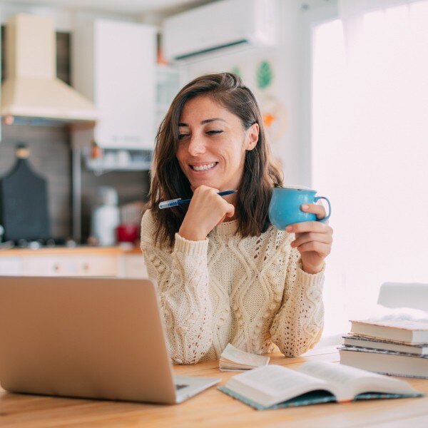 woman sitting at kitchen table holding a cup working laptop books