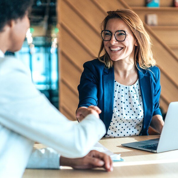 two coworkers sitting together at desk with laptop and notebook shaking hands smiling