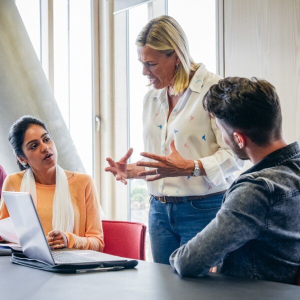 two coworkers sitting at desk one co worker standing facing each other talking