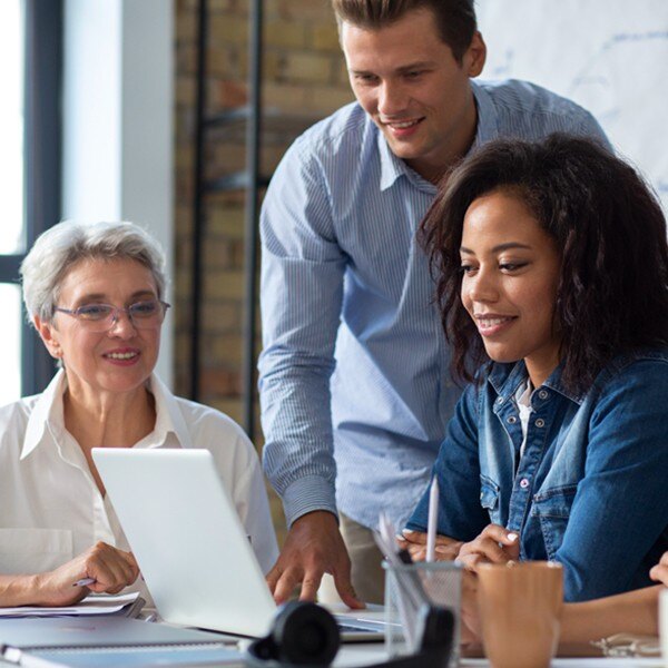 three coworkers sitting at desk in office one coworker standing looking at laptop