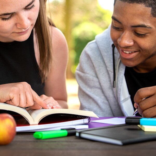 two students sitting next to each other outside reading book writing notes