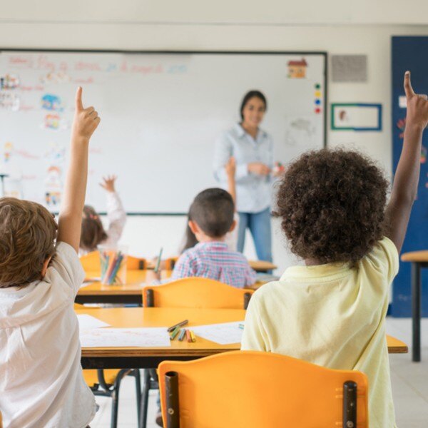 students sitting at desks raising hands teacher standing front of classroom smiling