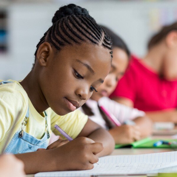 three students sitting at desks in classroom writing in notebook