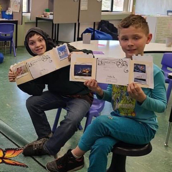 two students sitting in chairs in classroom holding up piece of paper smiling