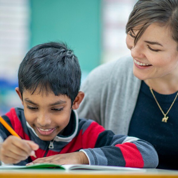 teacher and student sitting at desk in classroom writing in notebook