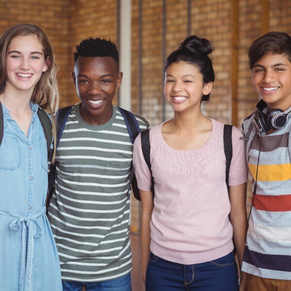 four students standing side by side in school hallway holding backpacks smiling