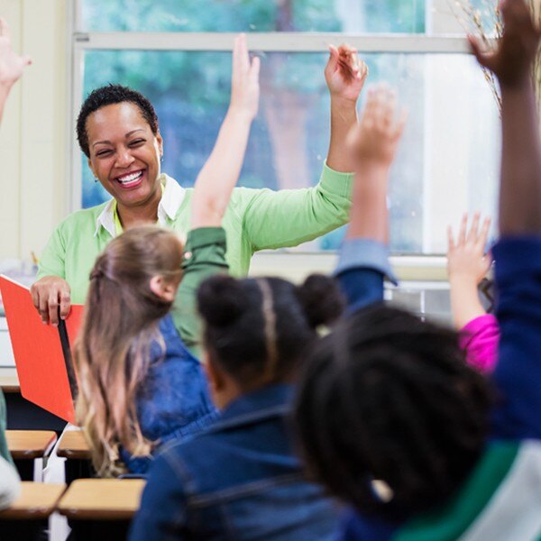 teacher sitting front of classroom smiling reading book to students sitting in desks raising hands