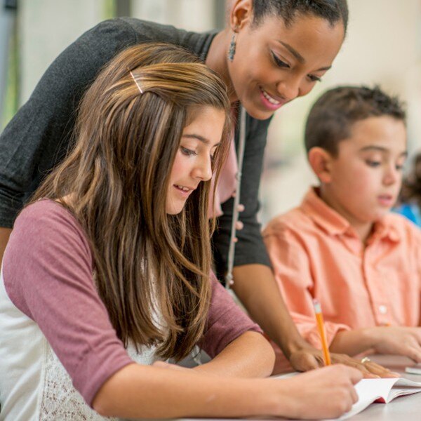 students sitting in classroom writing in notebooks teacher smiling helping