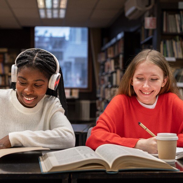 two students sitting at desk in library reading and writing in books